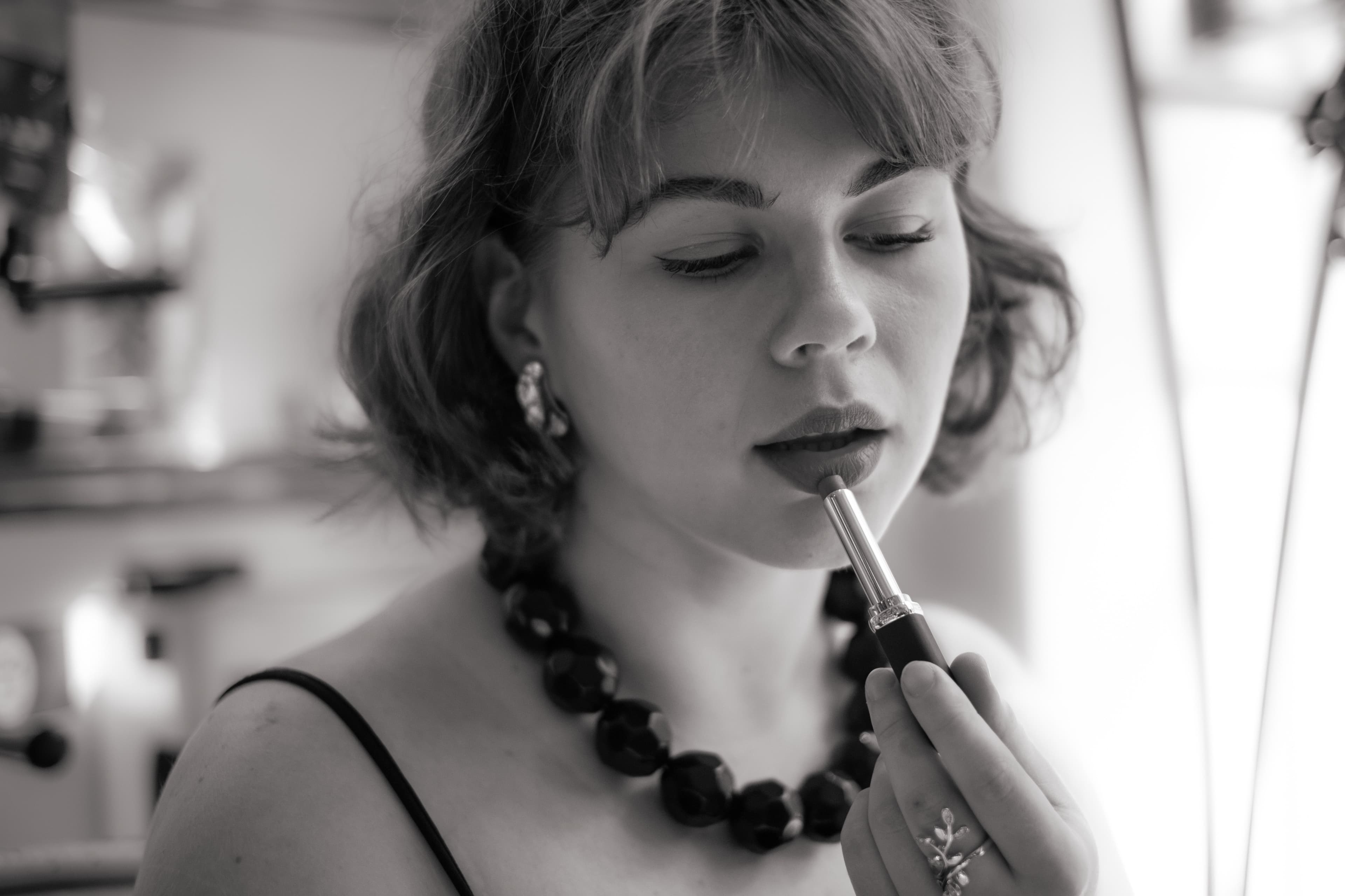 Retrato editorial en blanco y negro de una mujer con collar oscuro aplicándose labial frente a un tocador